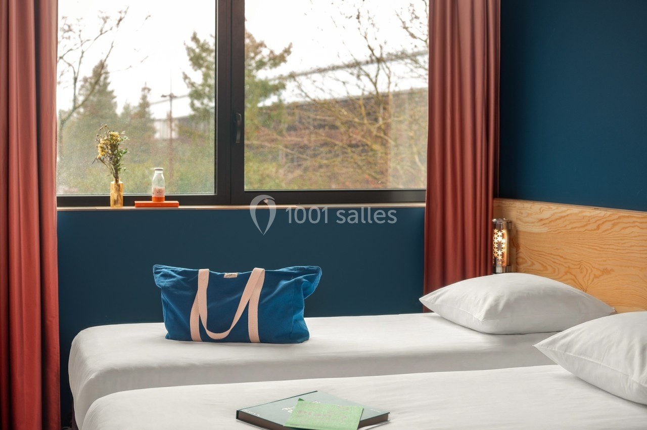 Deux lits simples avec draps blancs, sac bleu posé dessus, fenêtre avec rideaux rouges et vue sur des arbres.