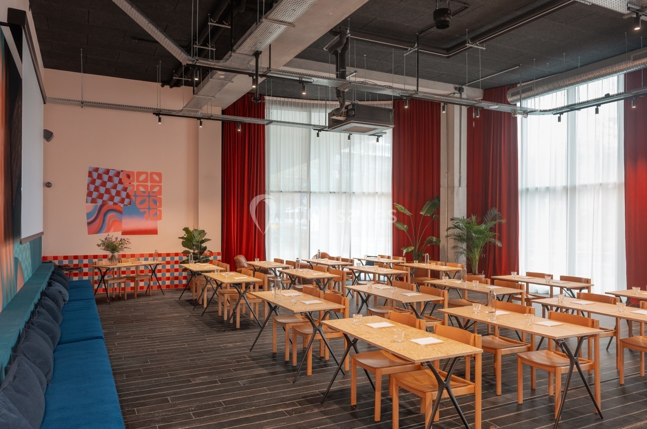 Salle lumineuse avec tables en bois, chaises, banquette bleue, rideaux rouges et décorations murales colorées.