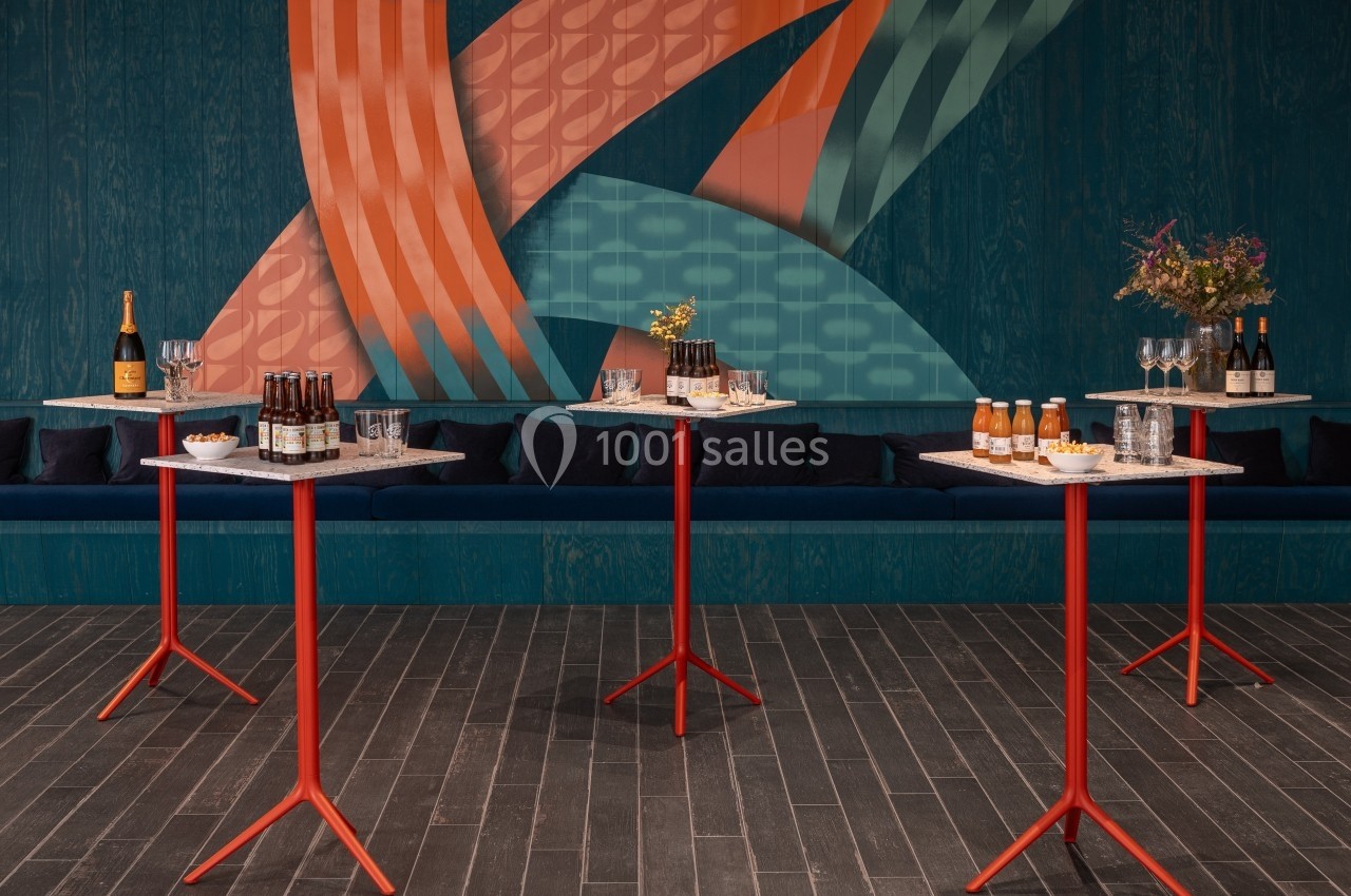 Tables hautes rouges avec boissons et amuse-bouches, devant un mur décoré de motifs colorés.