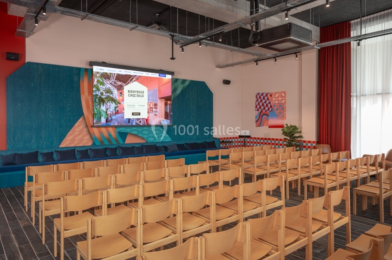 Salle de conférence lumineuse avec des rangées de chaises en bois, un écran de projection et une décoration colorée.