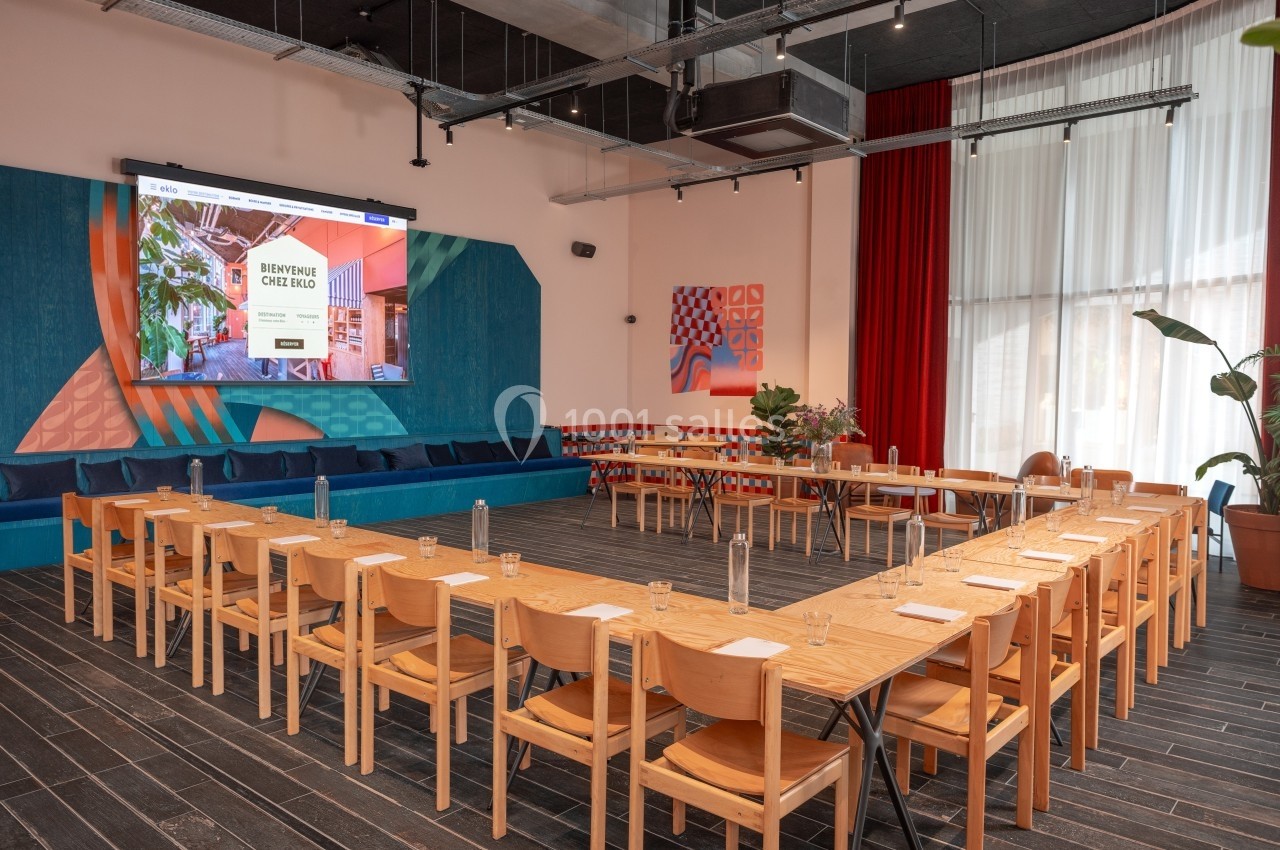 Salle de réunion lumineuse avec tables en U, chaises en bois, écran de projection et décoration colorée.
