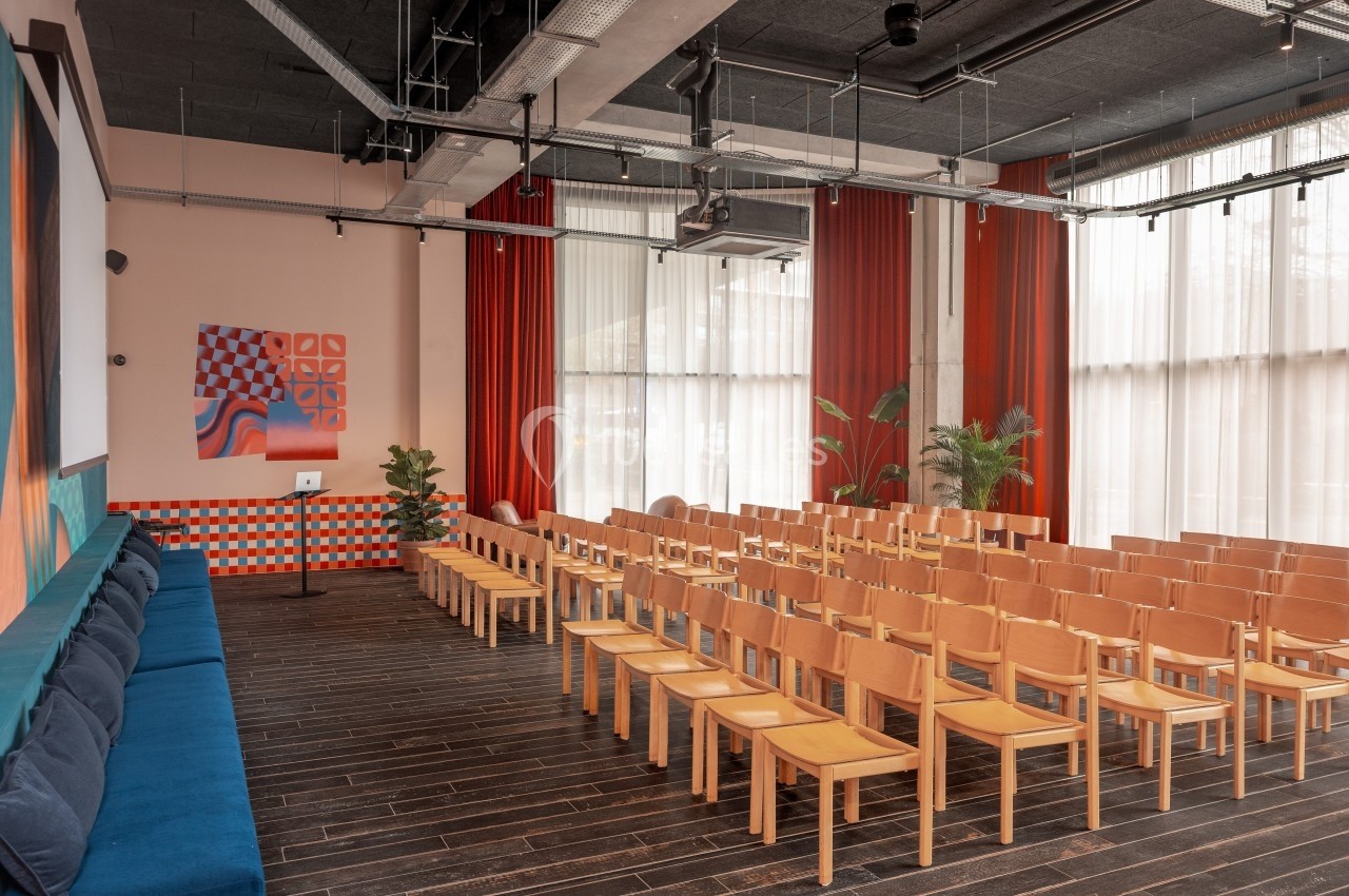 Salle de conférence lumineuse avec des rangées de chaises en bois, un podium et des décorations murales colorées.