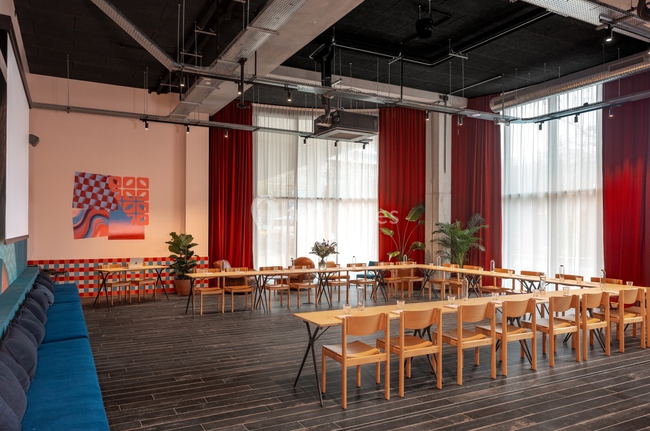 Salle lumineuse avec grandes fenêtres, rideaux rouges, chaises en bois, tables disposées en U et décorations murales…