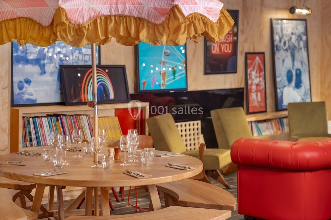 Table ronde en bois dressée avec vaisselle, entourée de bancs et fauteuils, sous un parasol coloré, dans une pièce décorée.
