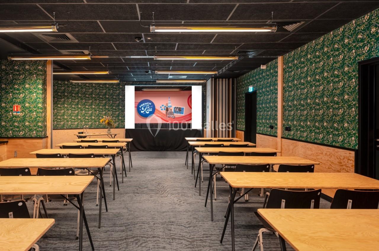 Salle de conférence avec des tables et chaises alignées, écran de projection allumé, décorations murales vertes.