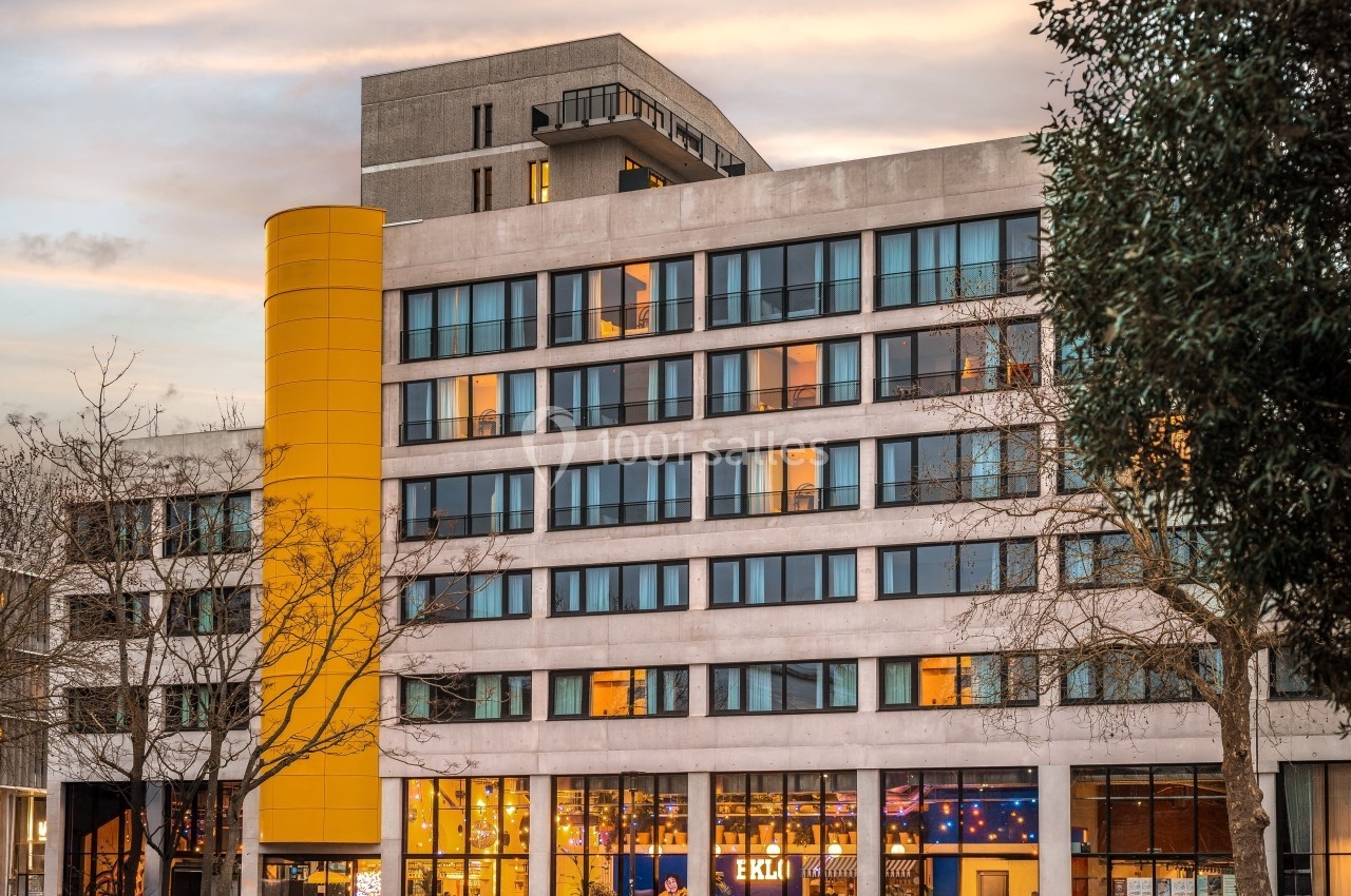 Façade d'un bâtiment moderne avec une colonne jaune, des fenêtres éclairées et des arbres en premier plan.