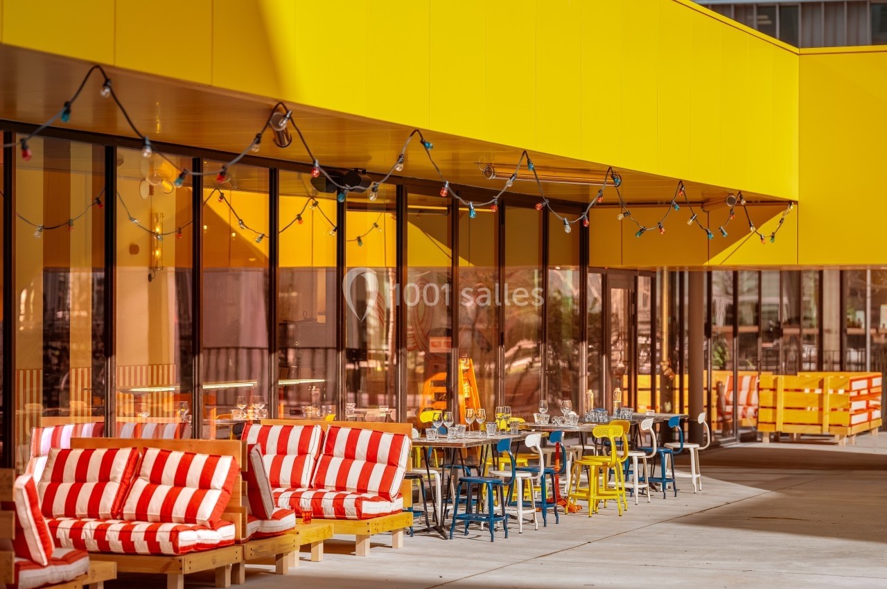 Façade moderne avec terrasse aménagée, mobilier coloré et guirlandes lumineuses sous un auvent jaune.