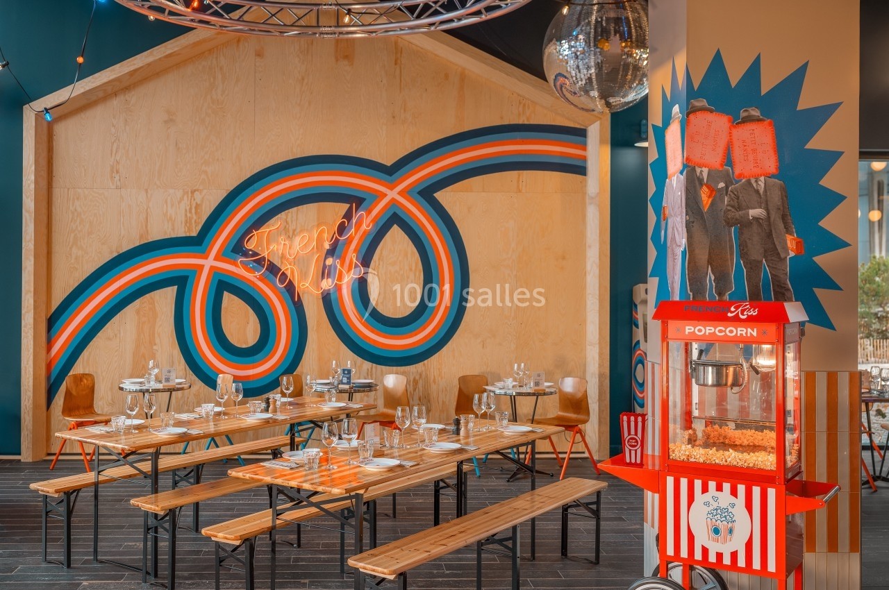 Salle de restaurant colorée avec tables en bois, machine à popcorn rouge et enseigne néon ’French Class’ sur un mur décoré.