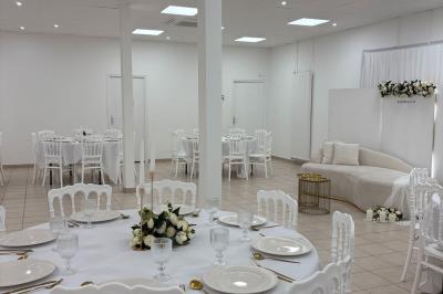 Salle de réception lumineuse avec tables rondes dressées, chaises blanches et décorations florales élégantes.