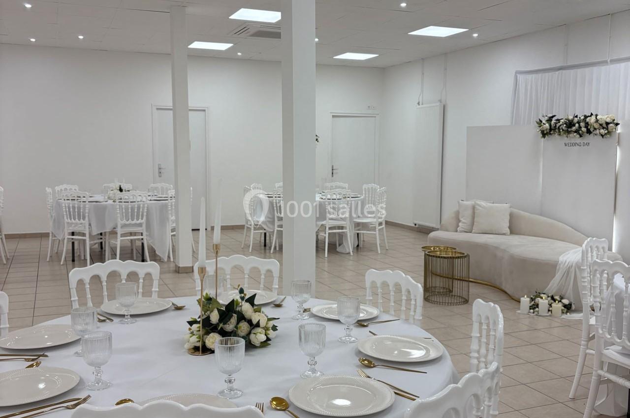 Salle de réception lumineuse avec tables rondes dressées, décorations florales blanches et espace lounge élégant.