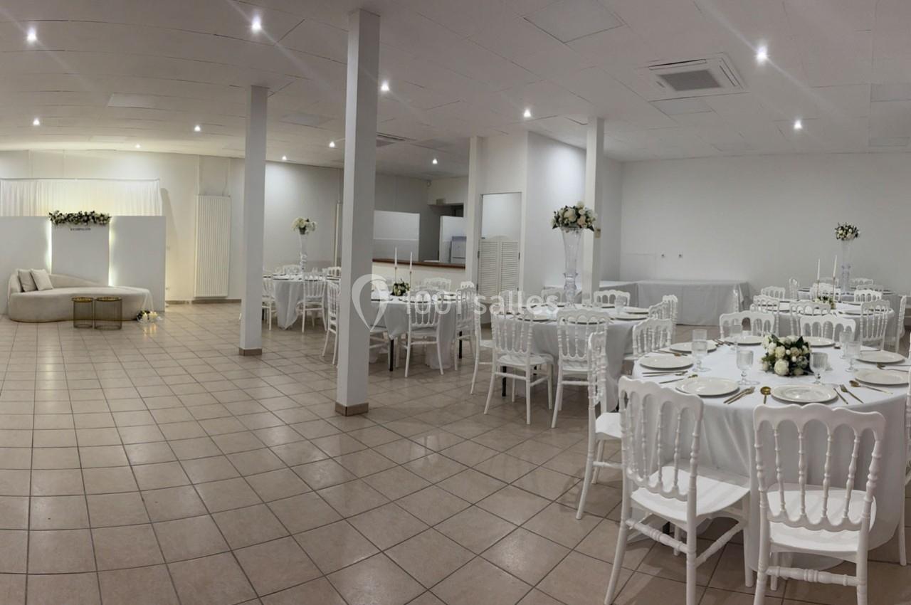 Salle de réception lumineuse avec tables rondes dressées, chaises blanches et décorations florales élégantes.