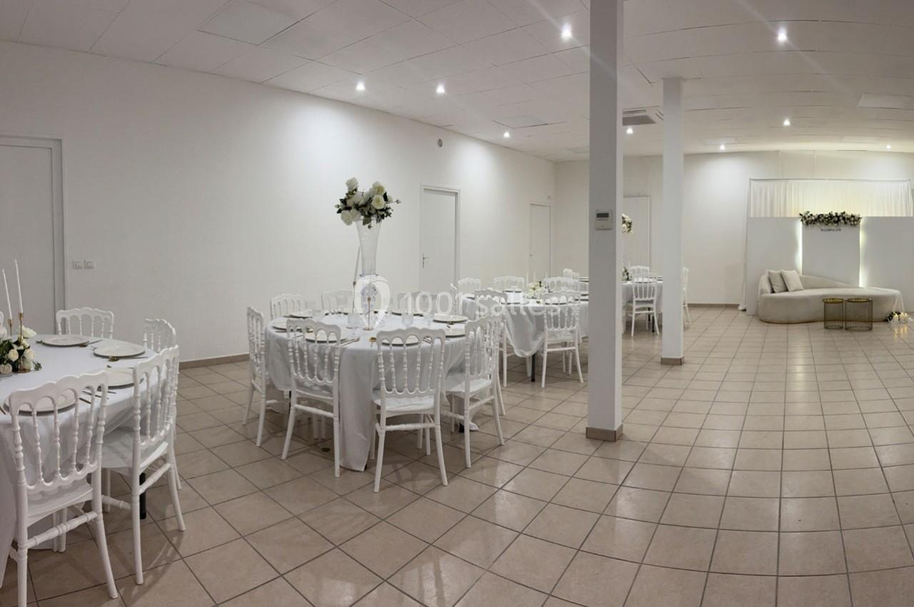 Salle de réception lumineuse avec tables rondes dressées, chaises blanches et décorations florales.