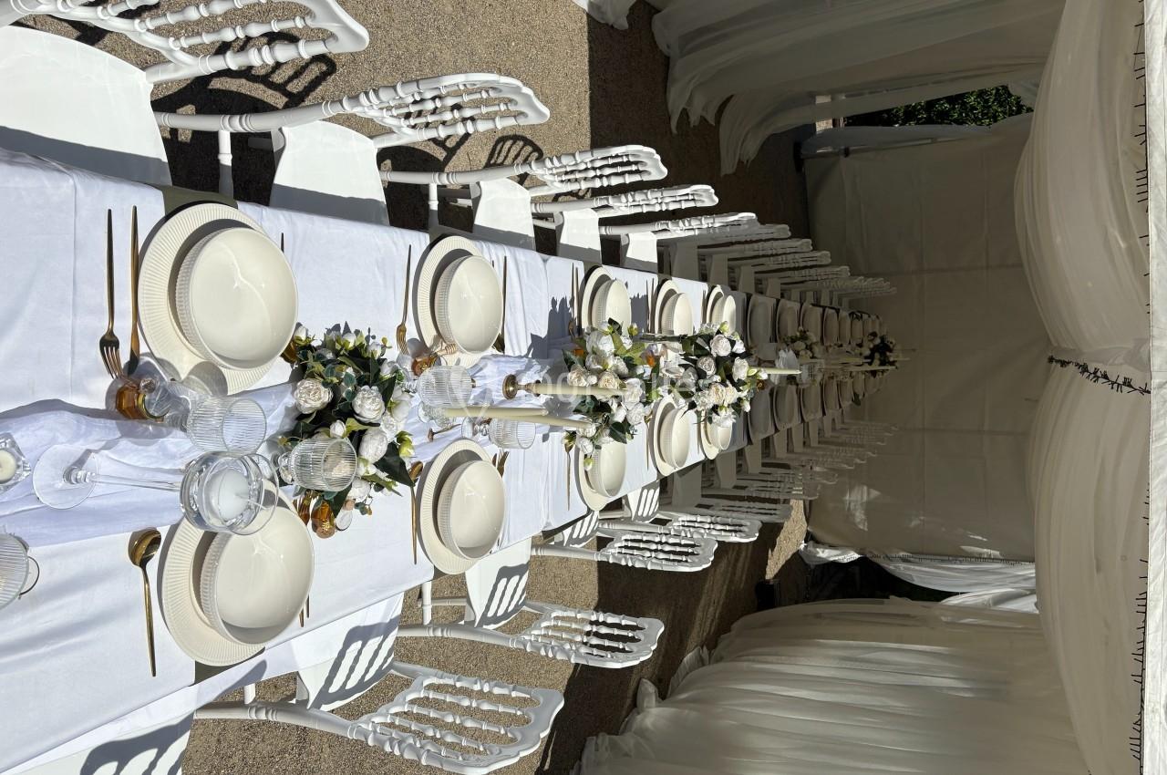 Table dressée pour un événement, avec nappes blanches, vaisselle élégante, fleurs et chaises alignées sous une tente.
