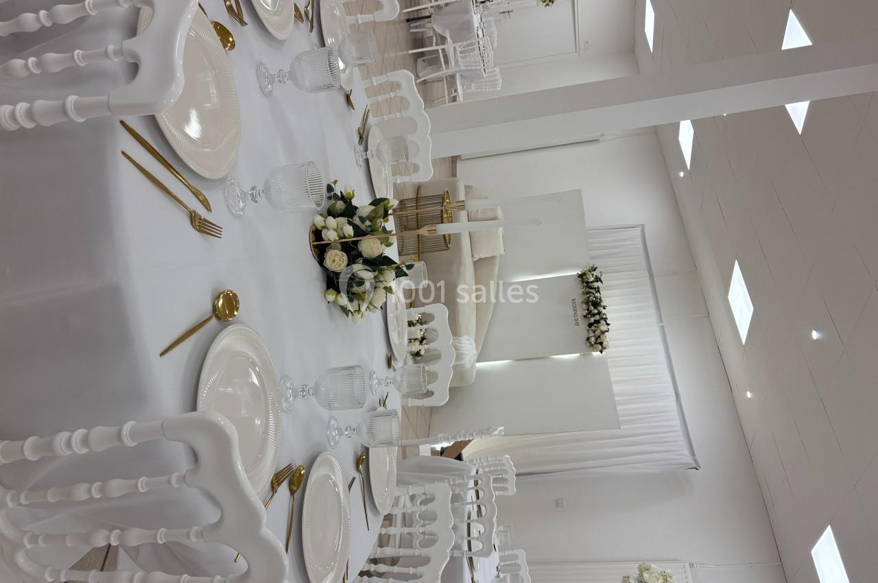 Salle de réception décorée en blanc avec tables dressées, chaises élégantes et arrangements floraux.