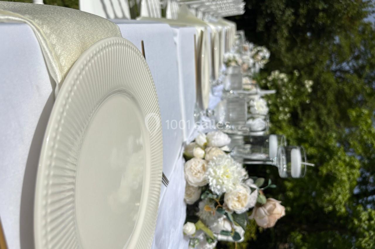 Table dressée en extérieur avec nappes blanches, vaisselle élégante et décorations florales dans un cadre verdoyant.