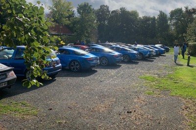 Plusieurs voitures blanches stationnées sur un parking en gravier, entouré de haies et de bâtiments.