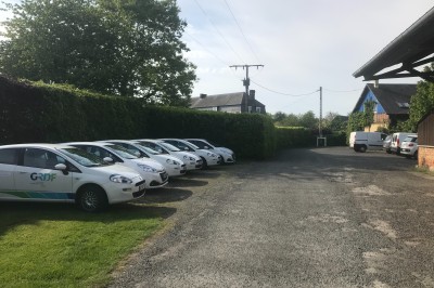 Plusieurs voitures blanches stationnées sur un parking en gravier, entouré de haies et de bâtiments.