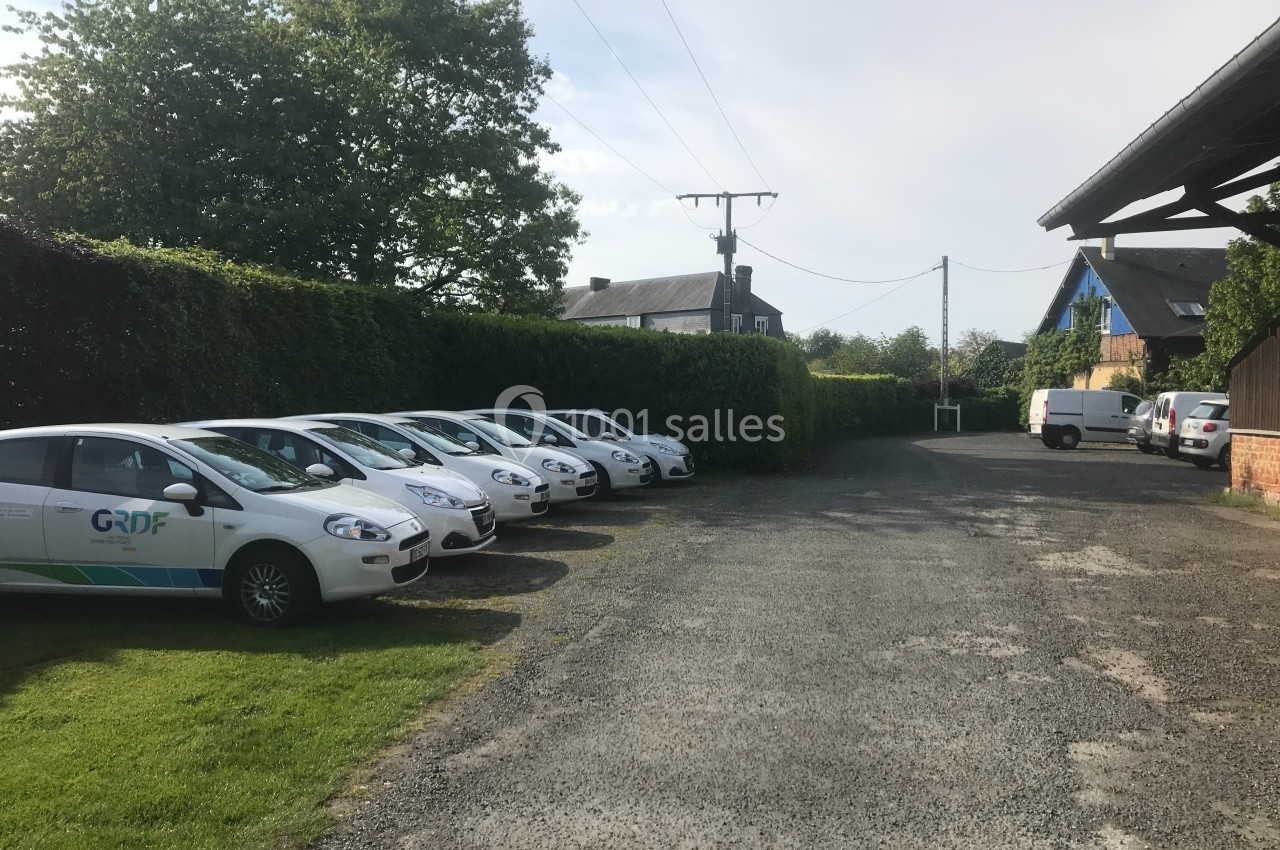 Plusieurs voitures blanches stationnées sur un parking en gravier, entouré de haies et de bâtiments.