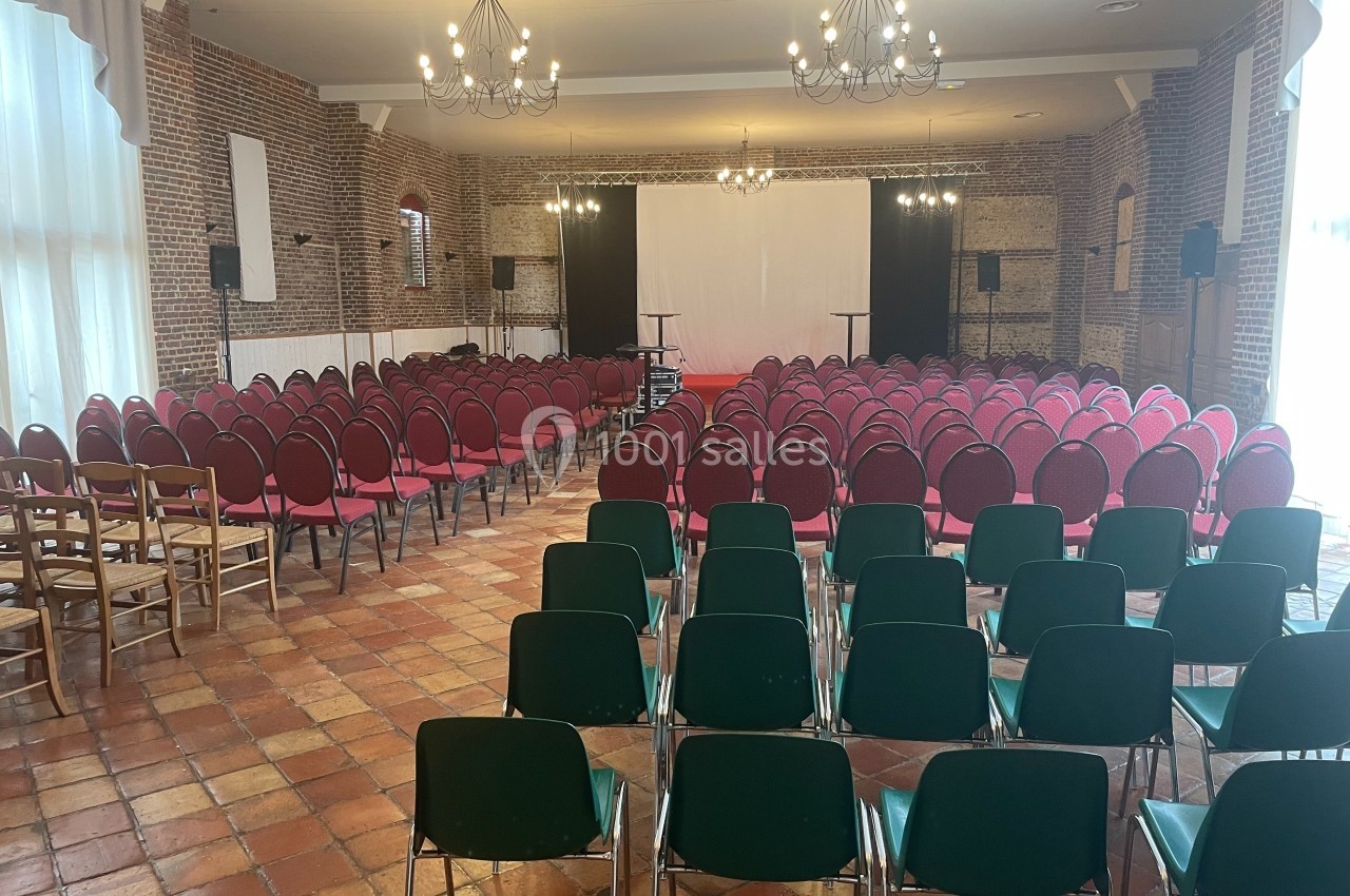 Salle de conférence avec des rangées de chaises rouges et vertes, éclairée par des lustres et un écran blanc en fond.