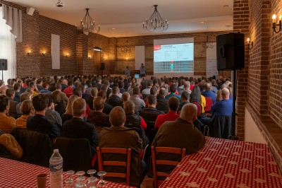 Plusieurs voitures blanches stationnées sur un parking en gravier, entouré de haies et de bâtiments.
