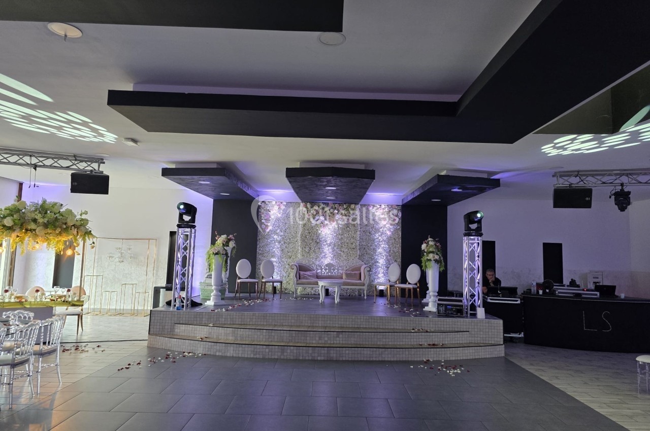 Salle de réception décorée avec une scène centrale, des fleurs, des éclairages et un espace pour les invités.