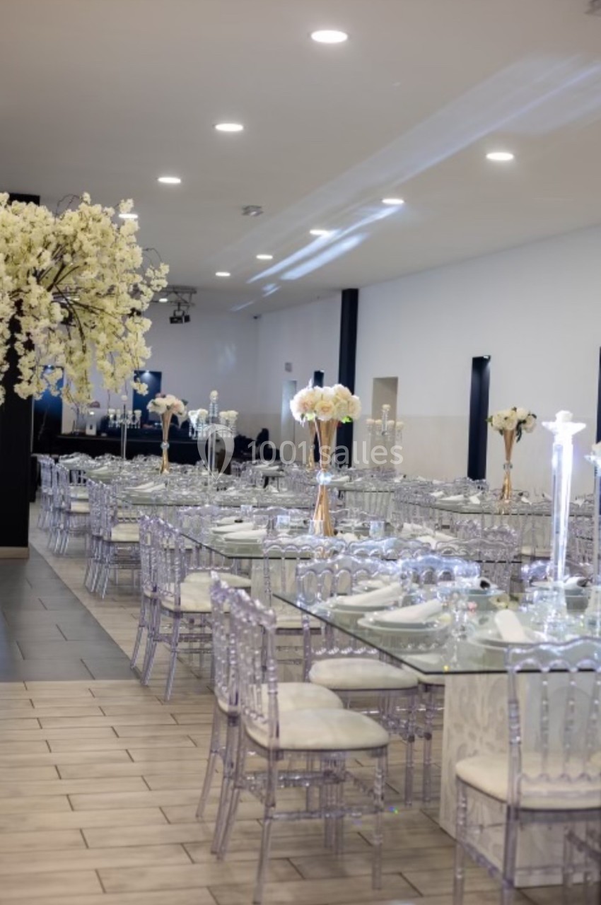 Salle de réception élégante avec tables dressées, chaises transparentes et décorations florales blanches.