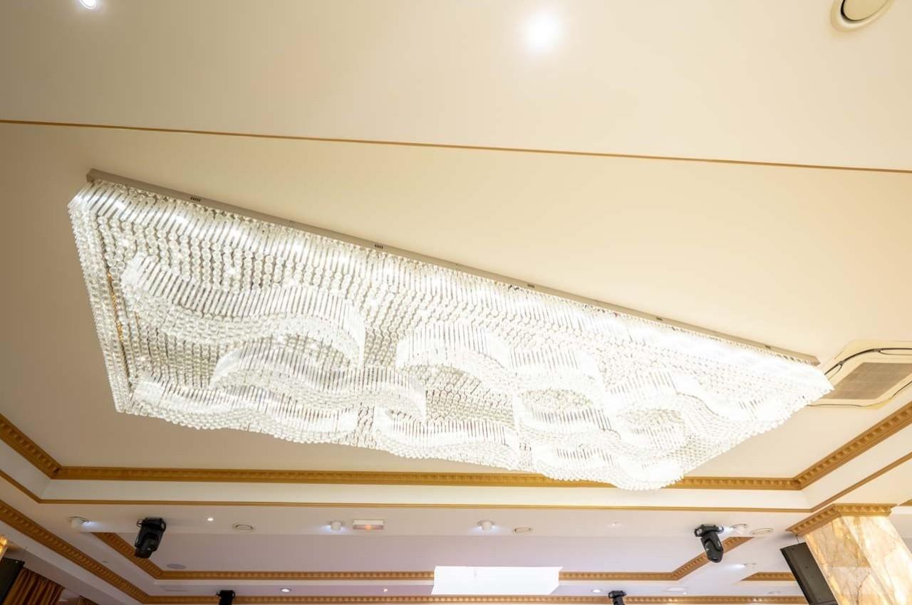 Plafonnier rectangulaire en cristal suspendu, éclairé, avec un design ondulé, dans une salle au plafond beige.