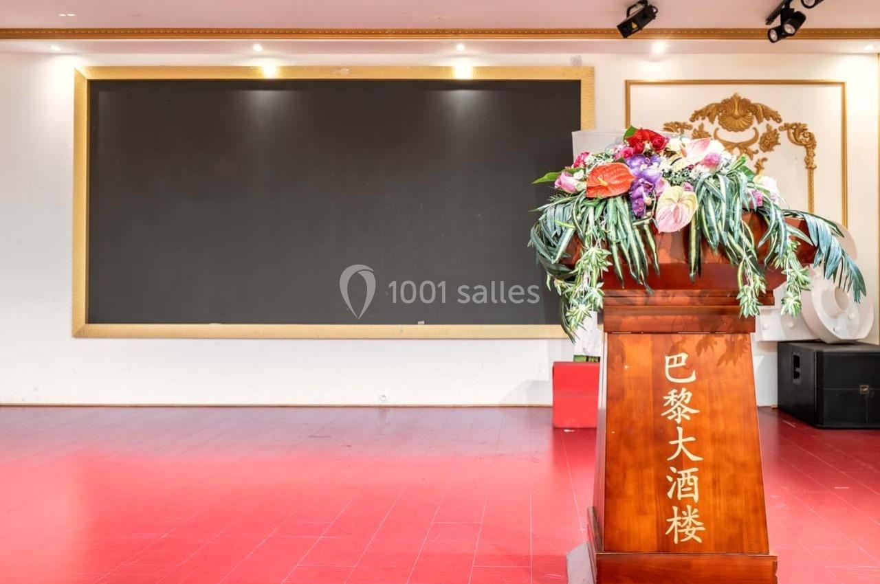 Un pupitre en bois décoré de fleurs devant un tableau noir dans une salle au sol rouge.