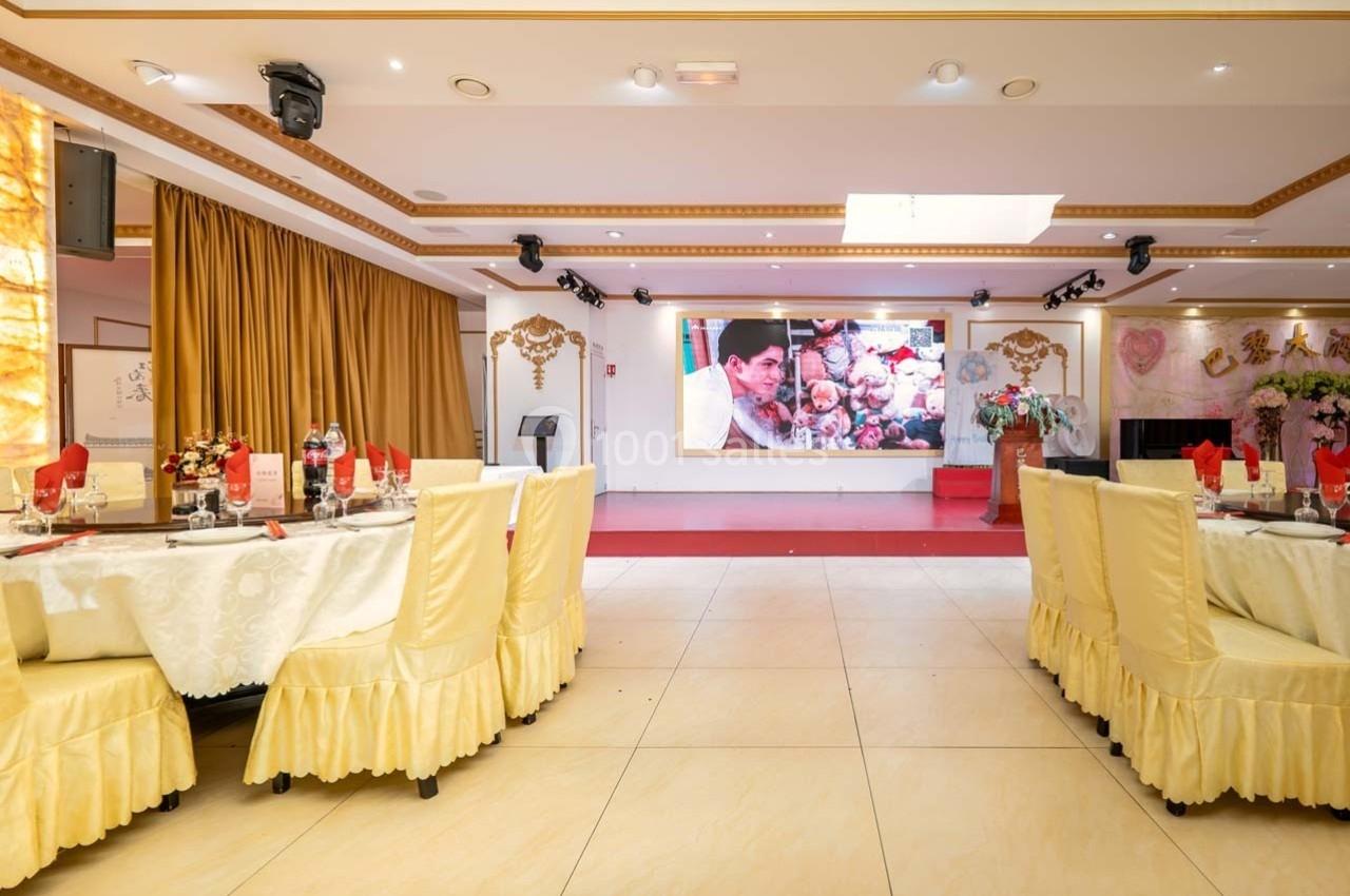 Salle de réception décorée avec des tables dressées, des chaises couvertes de housses jaunes et une scène ornée d'un écran…