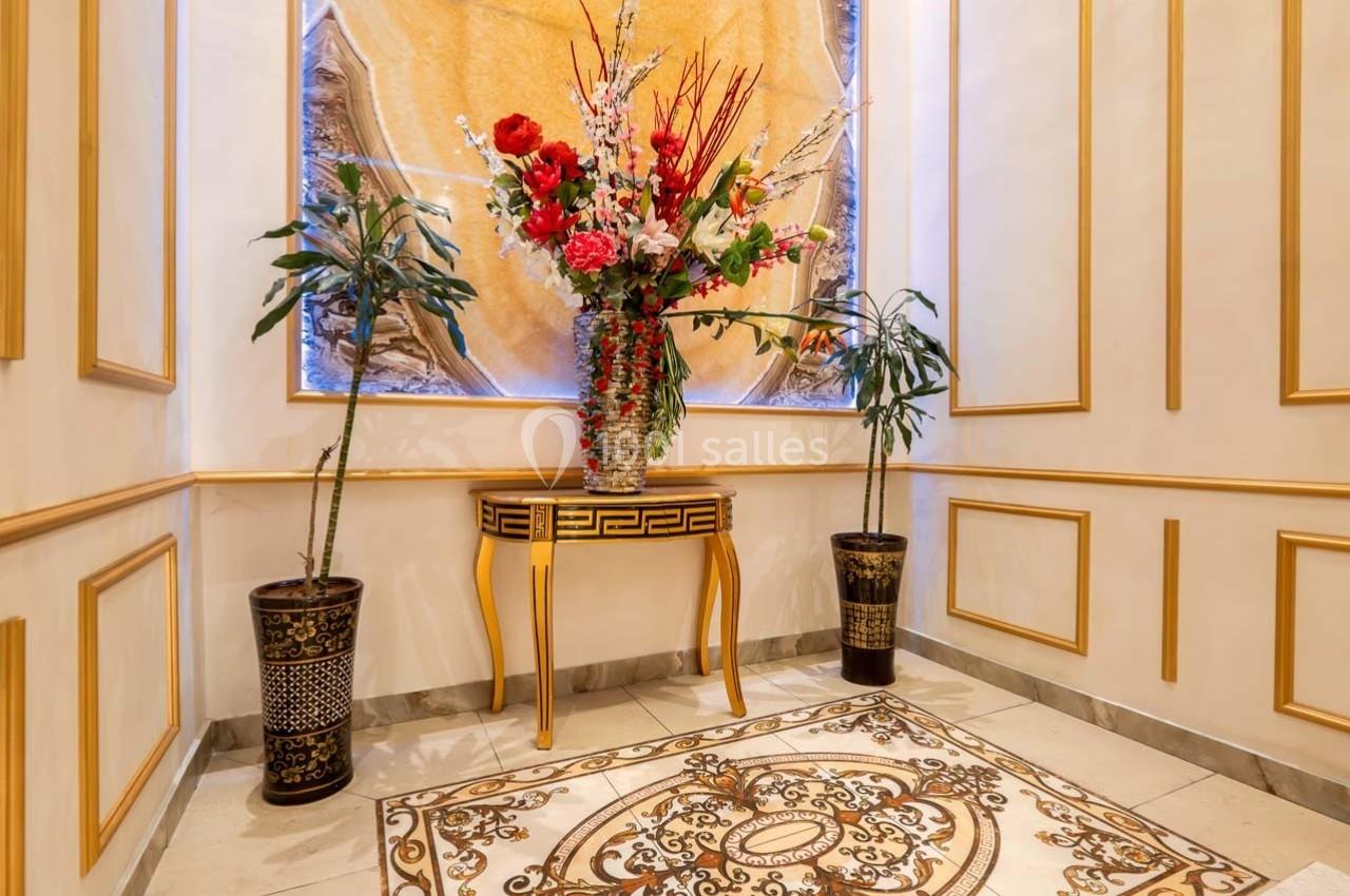 Vase de fleurs colorées sur une table ornée, encadré par deux plantes en pot dans un intérieur décoré de moulures dorées.