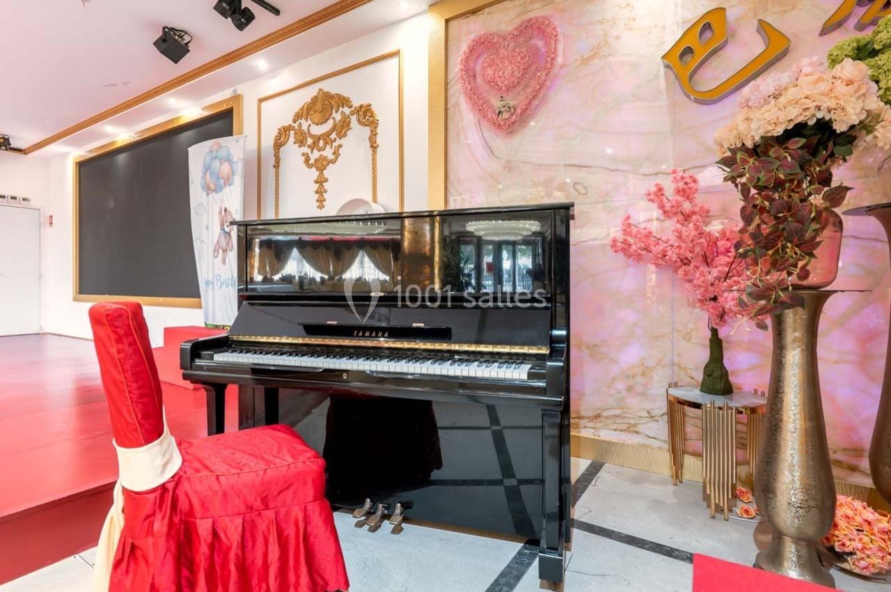 Piano droit noir devant une scène, entouré de décorations florales et d'éléments dorés sur un fond coloré.