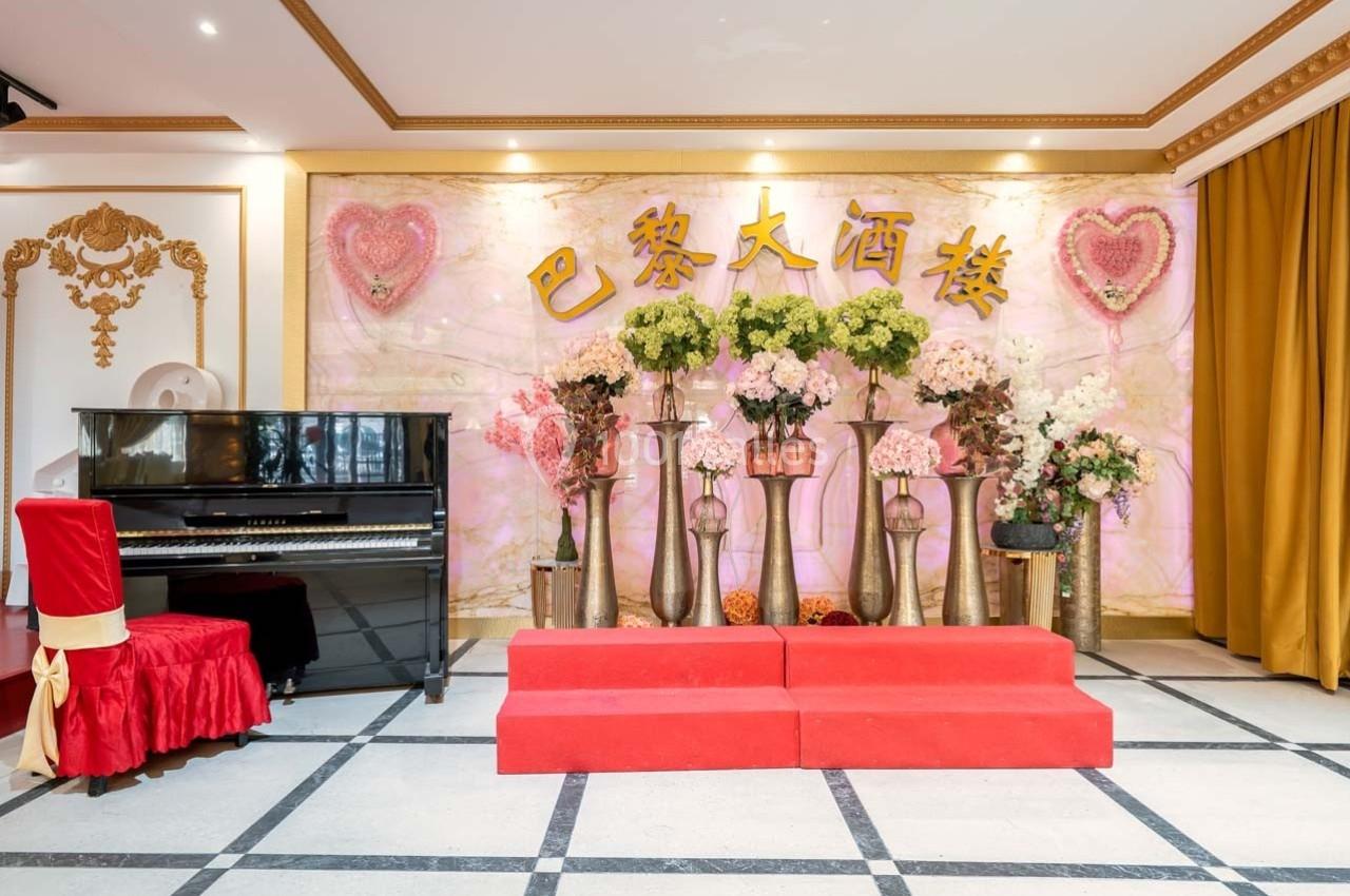 Salle décorée avec un piano noir, des fleurs dans des vases et une scène rouge devant un mur orné d'inscriptions chinoises.