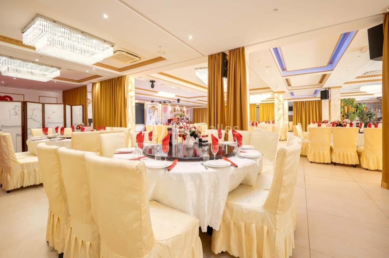 Salle de réception élégante avec tables rondes dressées, nappes blanches, serviettes rouges et décorations florales.