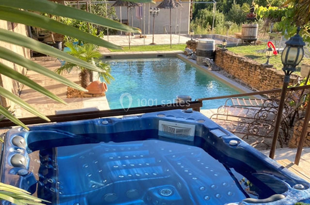 Vue d'un jacuzzi bleu au premier plan avec une piscine et un jardin arboré en arrière-plan.