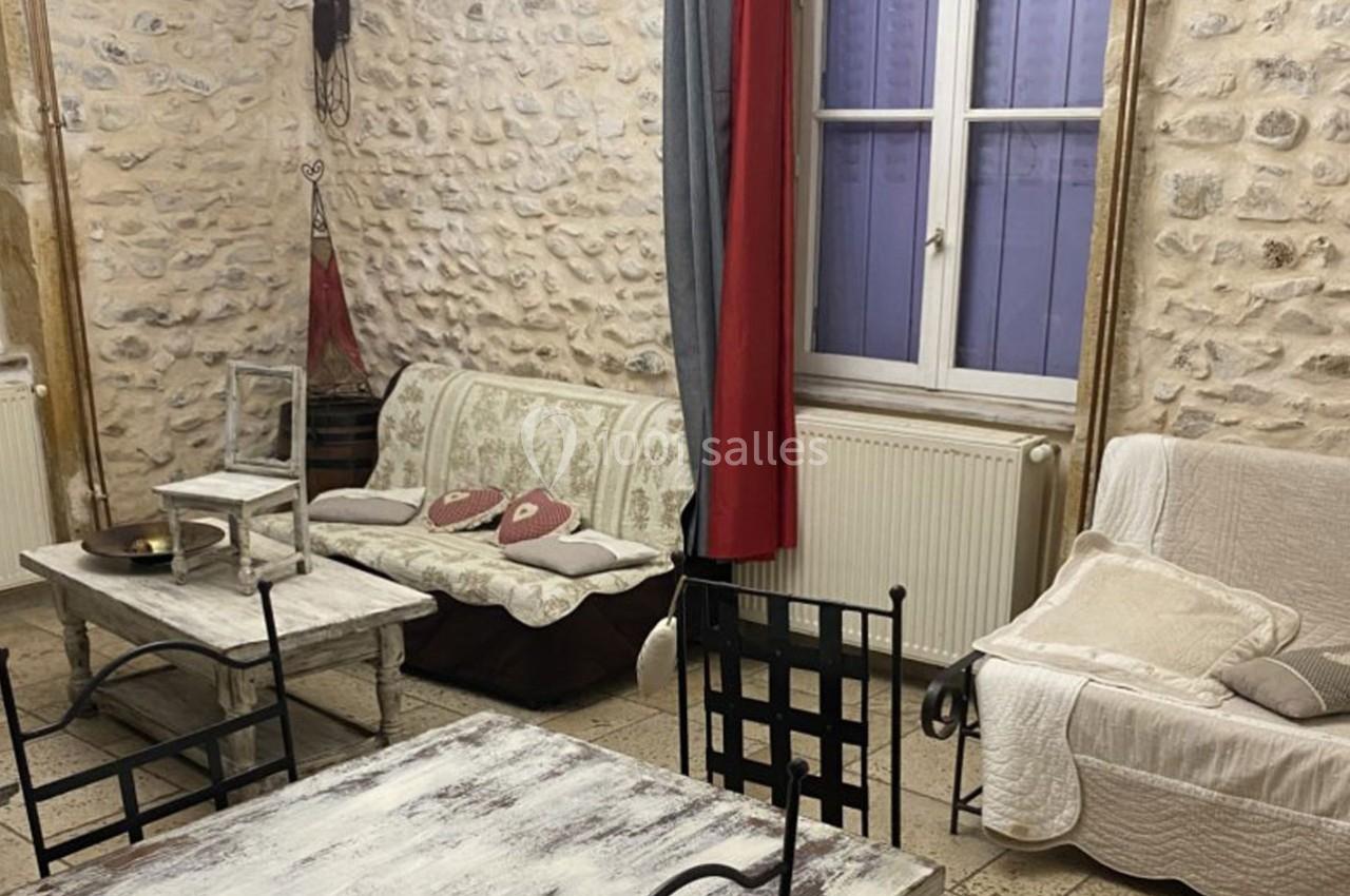 Salon rustique avec murs en pierre, mobilier en bois patiné, deux canapés, une table et une fenêtre avec rideaux.