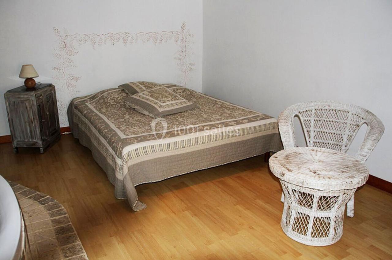 Chambre avec lit double, couvre-lit beige, table et chaise en osier blanc, sol en parquet clair et mur blanc décoré.