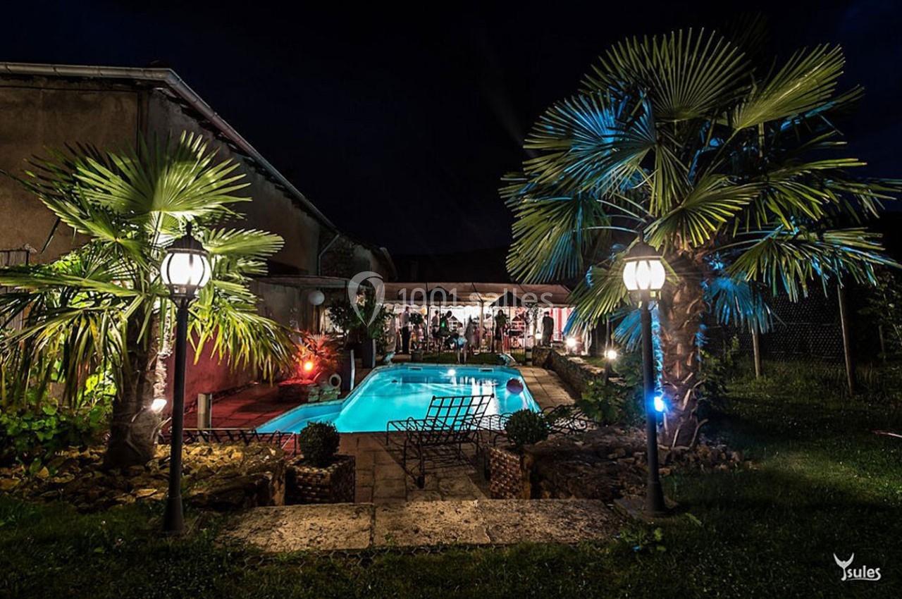 Jardin nocturne avec piscine éclairée, palmiers, lampadaires et terrasse animée en arrière-plan.