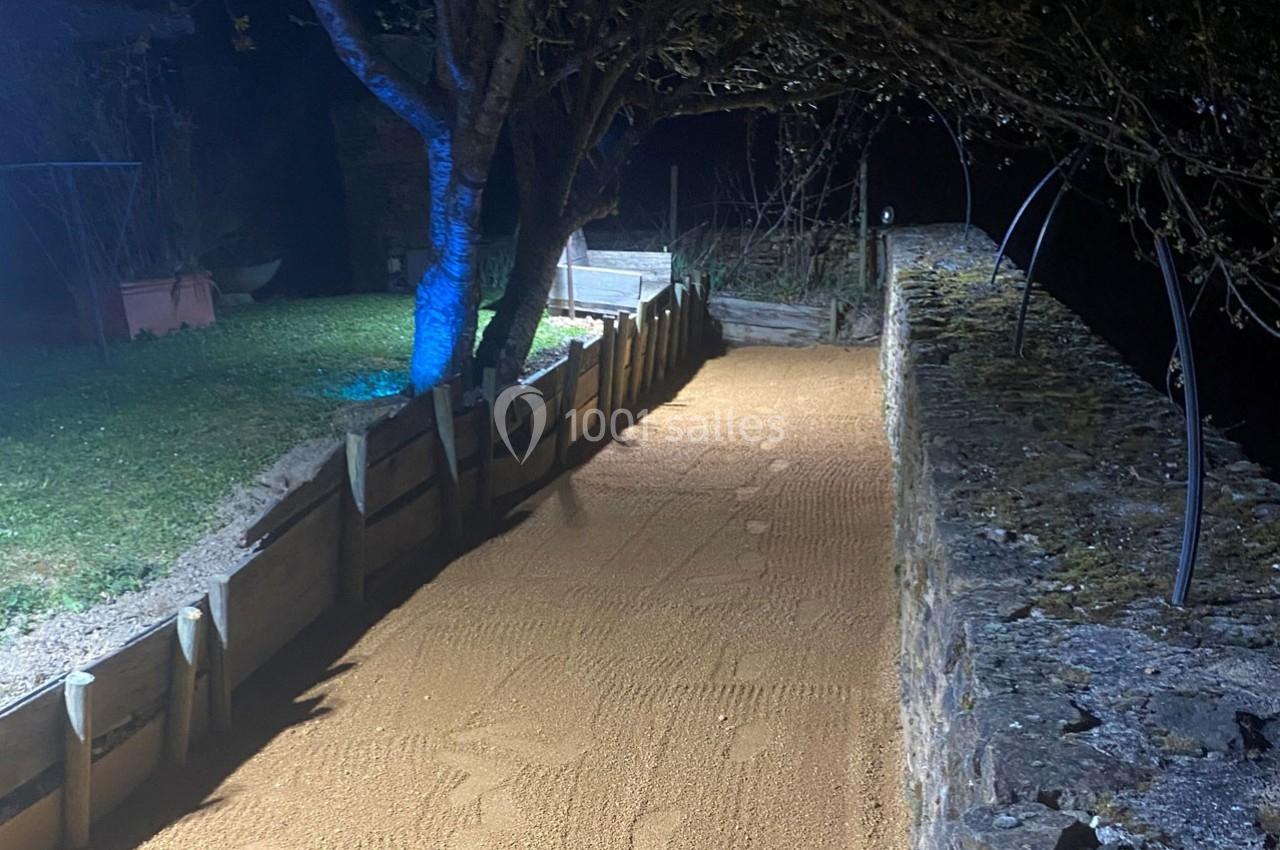 Allée en sable bordée d'un mur en pierre et de barrières en bois, éclairée par une lumière nocturne.