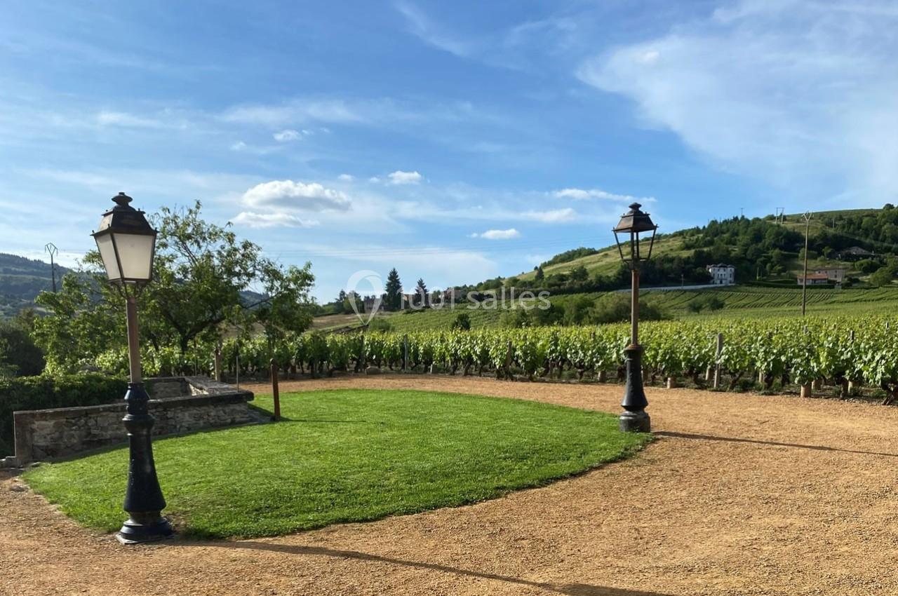 Pelouse entourée de chemins en gravier, lampadaires et vignobles en arrière-plan sous un ciel dégagé.