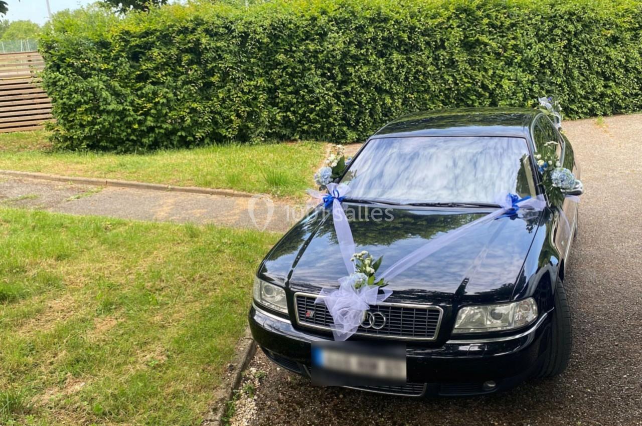Voiture noire décorée de rubans blancs et de fleurs blanches et bleues, stationnée près d'une haie verte.