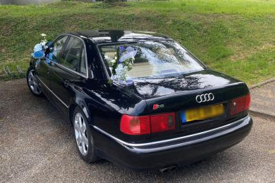 Voiture noire décorée de fleurs blanches et bleues, garée sur un parking près d'une pelouse.