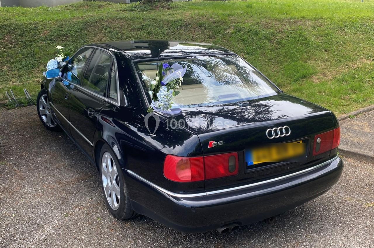 Voiture noire décorée de fleurs blanches et bleues, garée sur un parking près d'une pelouse.