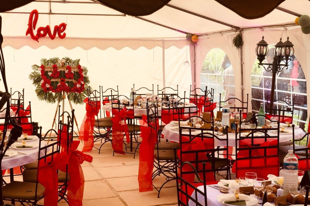 Salle de réception sous tente, décorée avec des chaises ornées de nœuds rouges et une enseigne lumineuse ’love’.