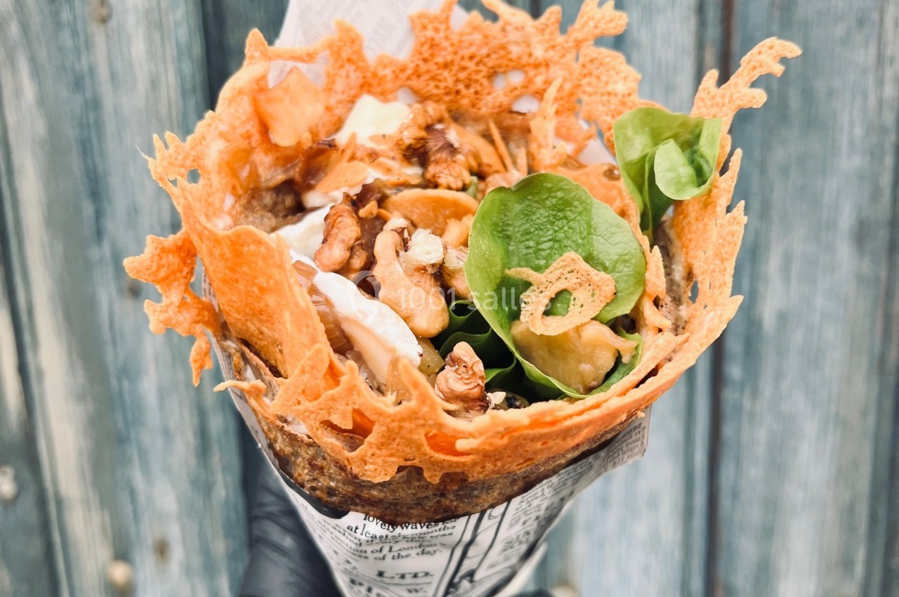 Crêpe garnie de légumes, noix, fromage croustillant et sauce, tenue dans une main gantée devant un fond en bois.