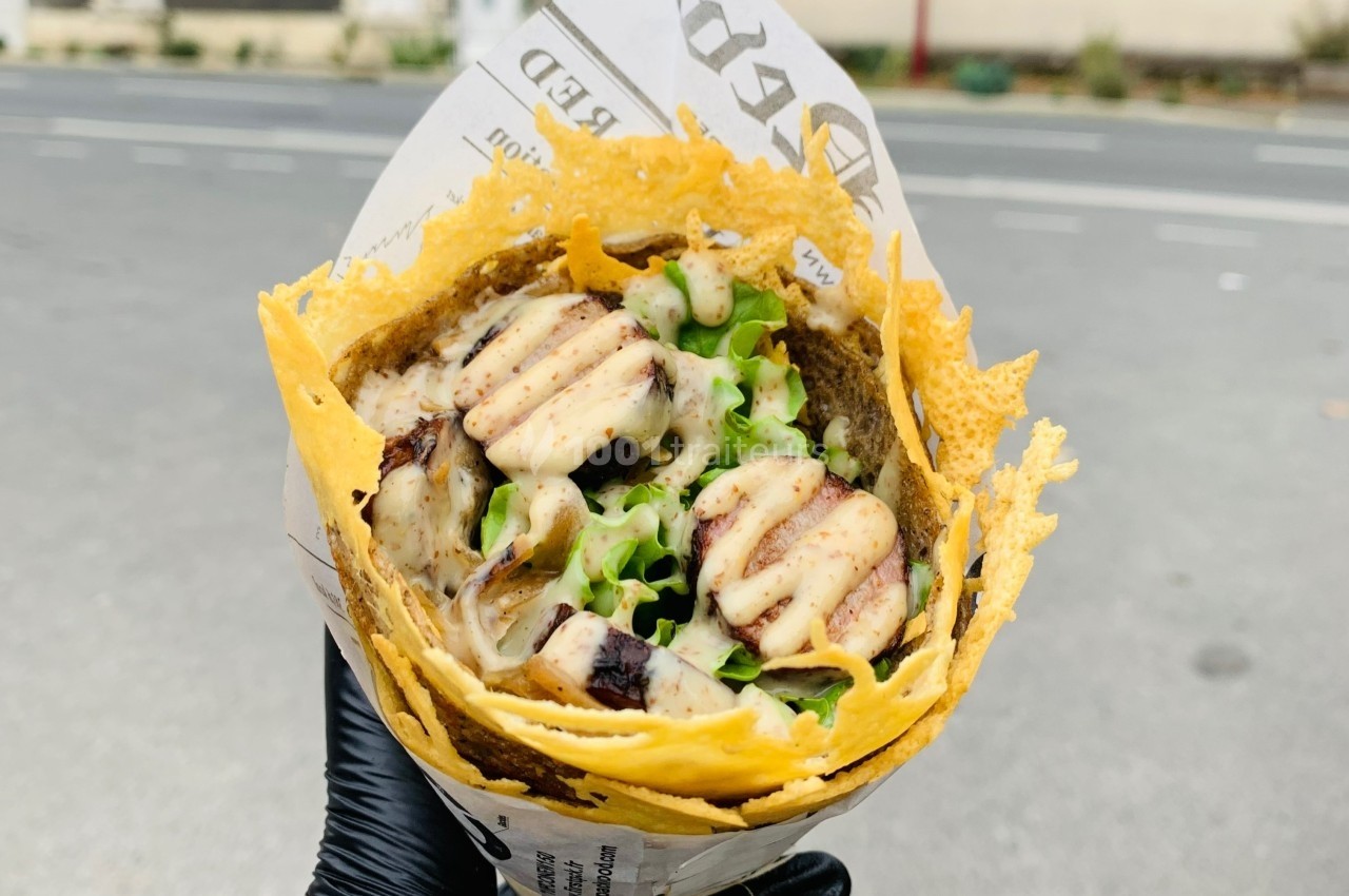 Wrap garni de poulet grillé, salade et sauce, enveloppé dans une crêpe au fromage croustillante, tenu par une main gantée.