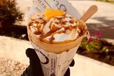 Crêpe garnie de crème fouettée, sauce caramel, éclats de noisettes et boule de praliné, tenue dans une main.