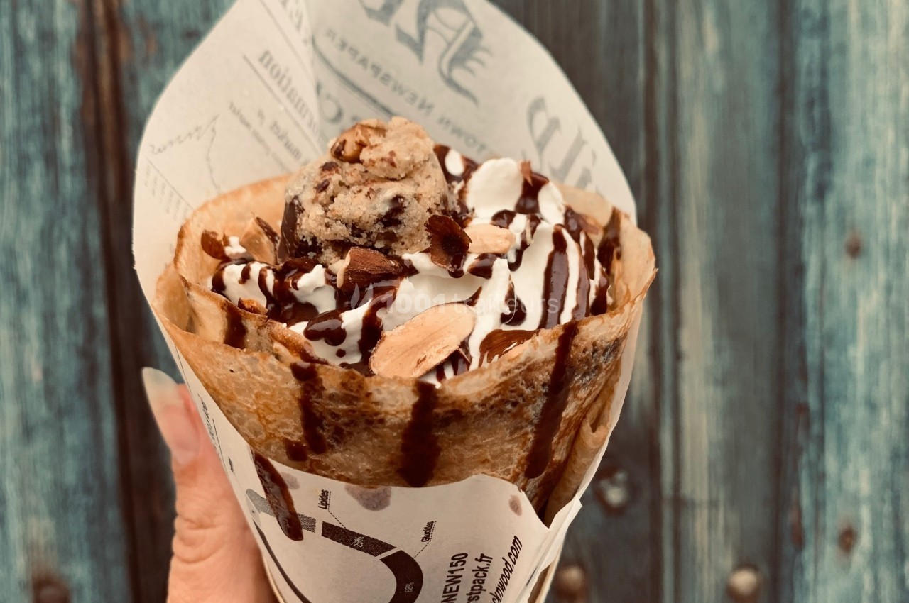 Crêpe garnie de glace, chantilly, amandes et sauce chocolat, présentée dans un papier imprimé devant un fond en bois.