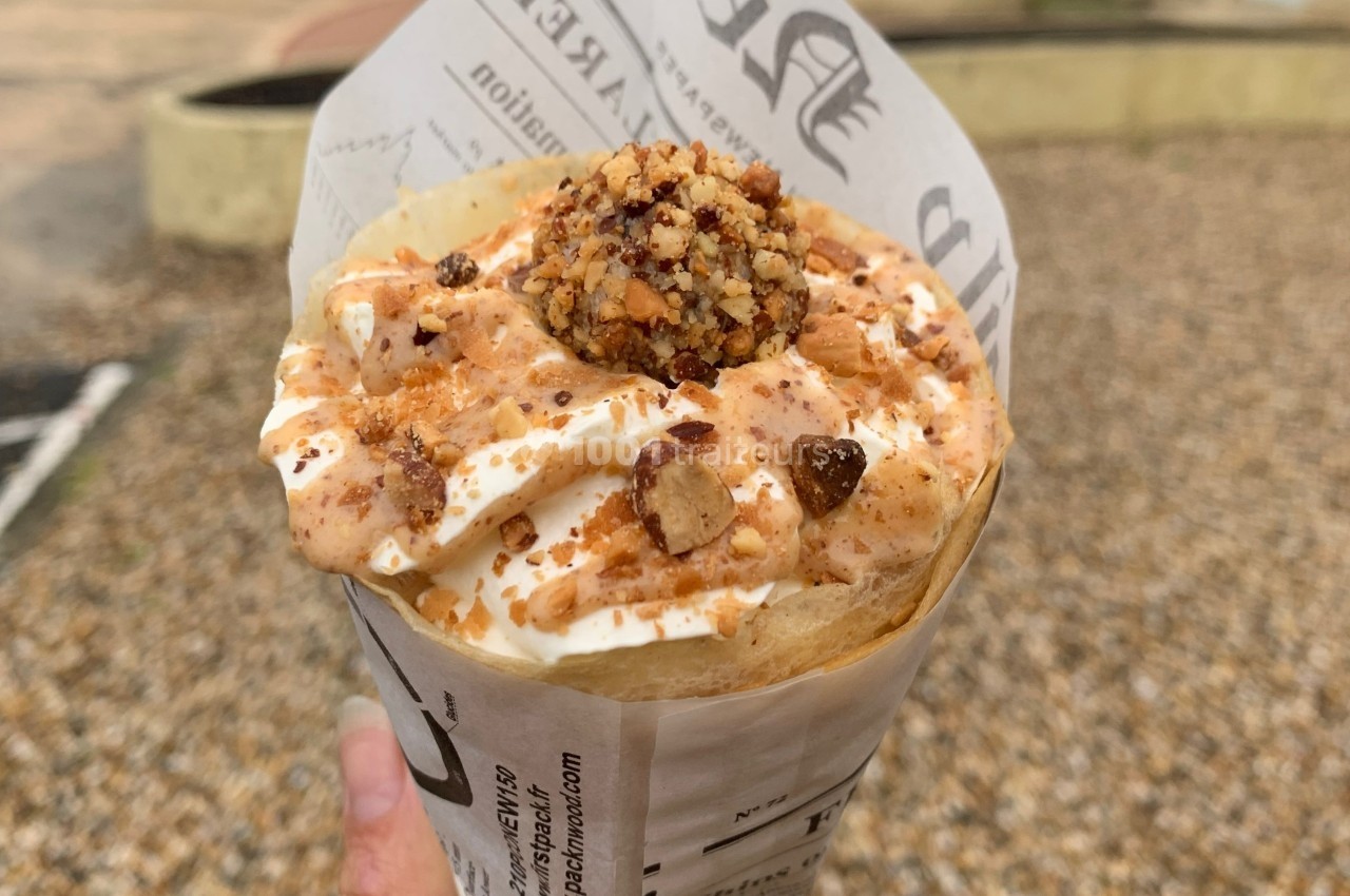 Crêpe garnie de crème fouettée, sauce caramel, éclats de noisettes et boule de praliné, tenue dans une main.