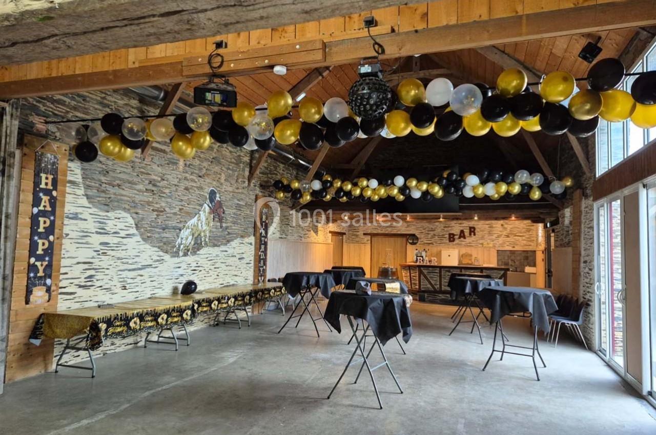 Salle décorée pour une fête avec des ballons noirs, dorés et blancs, tables hautes et un bar en fond.