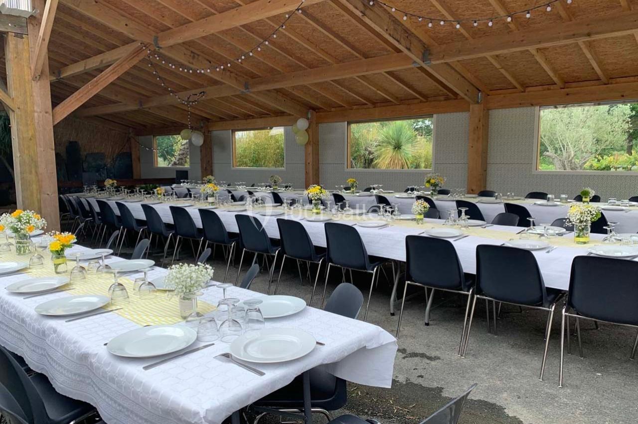 Tables longues dressées avec nappes blanches, vaisselle et bouquets de fleurs, sous un préau décoré de guirlandes lumineuses.